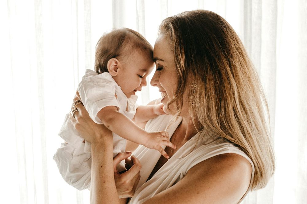 Mãe sorrindo com o bebê no colo perto da janela